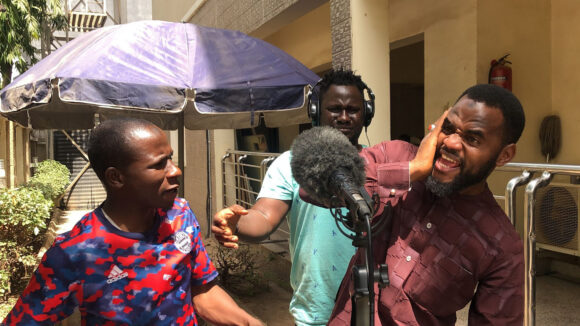 Three radio actors gathered around a microphone, wearing earphones and showing animated expressions.
